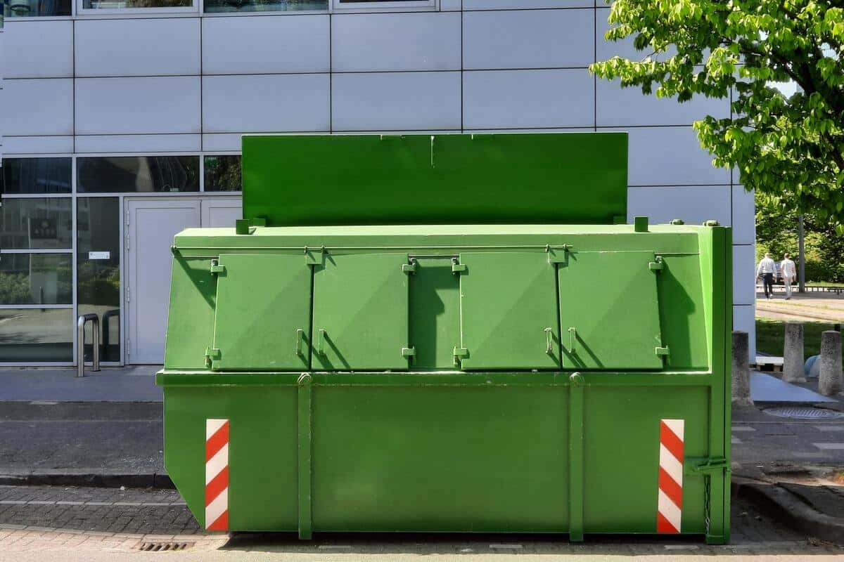 Benne verte à couvercles installée en bord de trottoir, devant un bâtiment et un arbre en feuilles