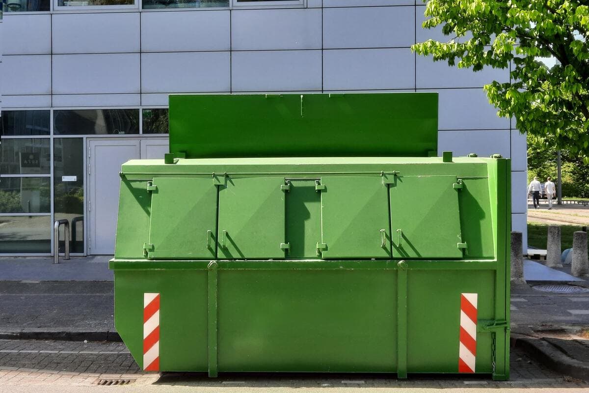 Benne verte à couvercles installée en bord de trottoir, devant un bâtiment et un arbre en feuilles